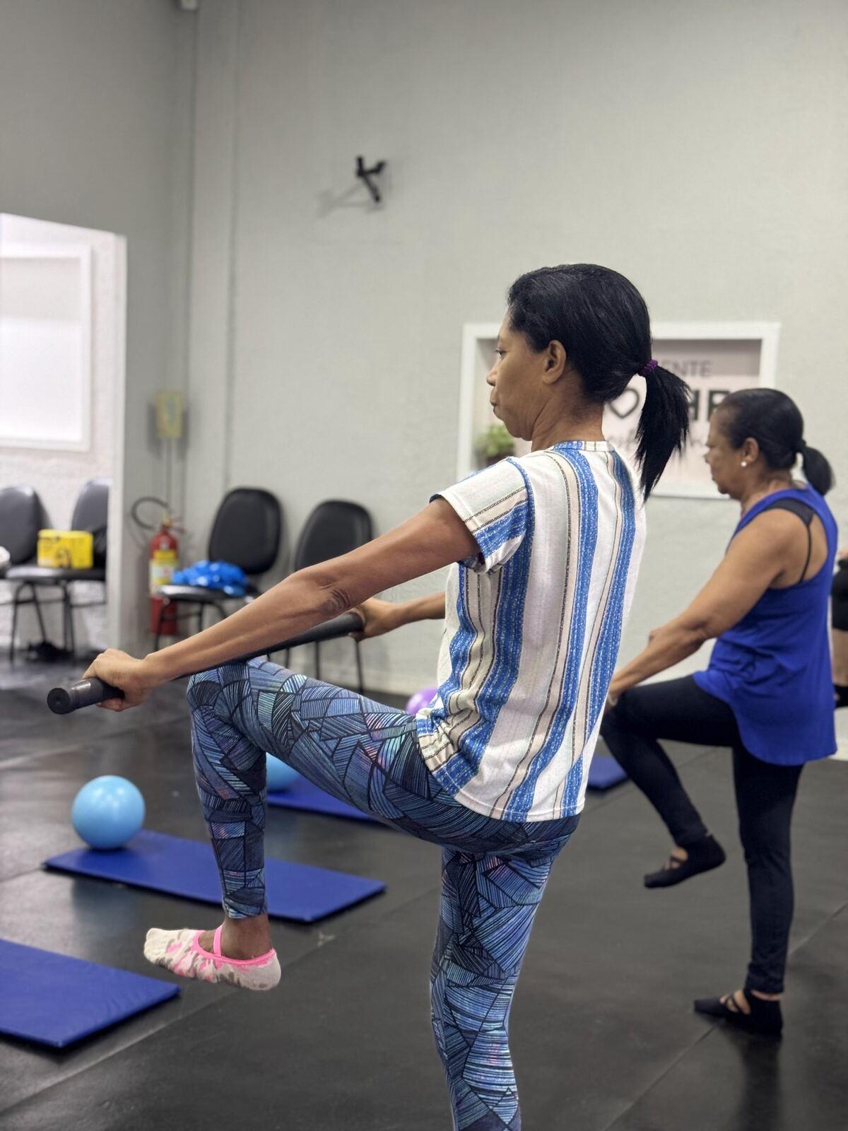 Aluna de Pilates levantando a perna com uma barra nas mãos, em uma sala com colchonetes
